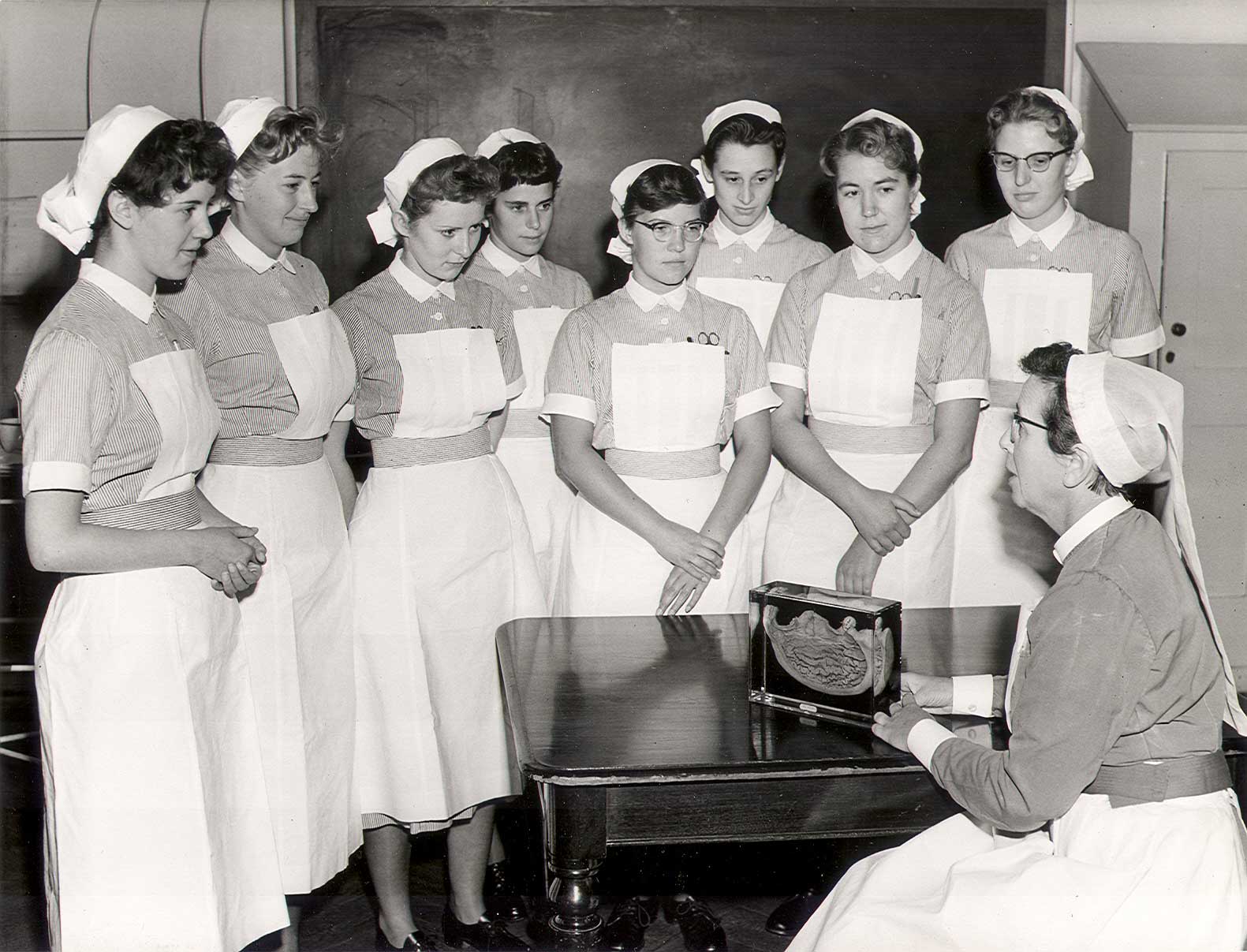 SBHX8/567 "Winifred Hector teaching student nurses in uniform, c.1960. Hector seated at right with gastric specimen on table. Eight second year student nurses [indicated by their belts] standing, from the Oct 1958 set." 1960 Nursing Mirror